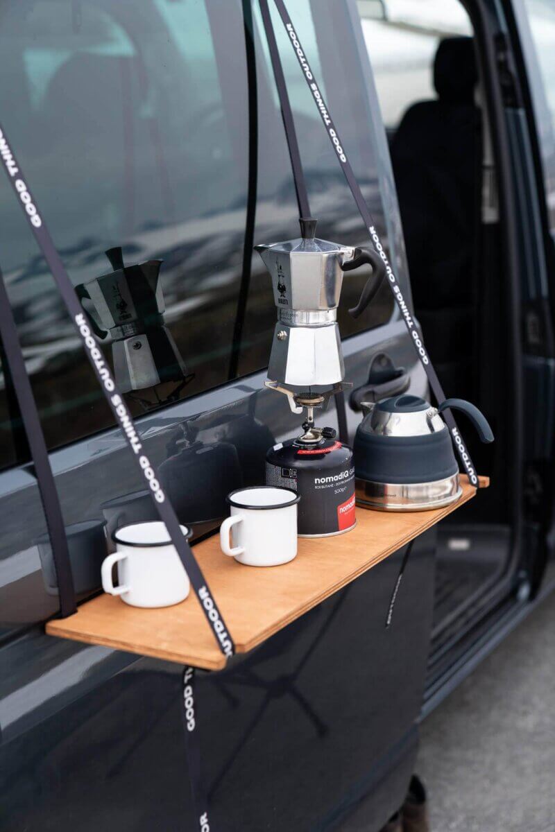 Ein Regalbrett ist an der Außenseite der Schiebetür angebracht. Auf dem Regal steht ein Gaskocher, eine Kaffeekanne und zwei Kaffeebecher.